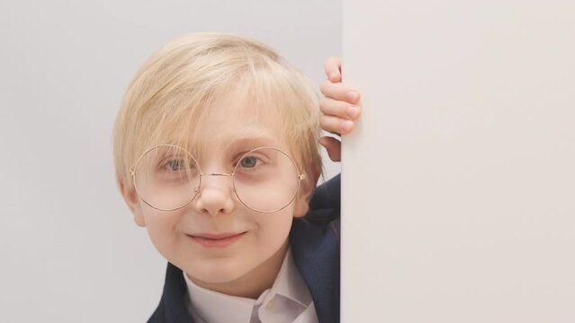 Curious smiling schoolboy in big glasses peeks out from behind the wall. Happy blond boy. Close-up video.