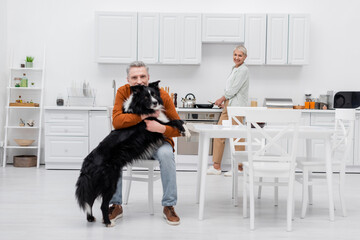 Smiling senior woman cooking and looking at husband with border collie in kitchen.