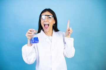 Young brunette woman wearing scientist uniform holding test tube over isolated blue background surprised and thinking with her finger on her head that she has an idea.