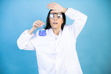 Naklejka premium Young brunette woman wearing scientist uniform holding test tube over isolated blue background Touching forehead for illness and fever, flu and cold, virus sick