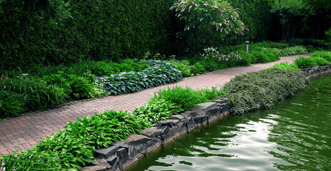 pond shore sidewalk with brick stone tiles in park with plants and decorative water pond filled...