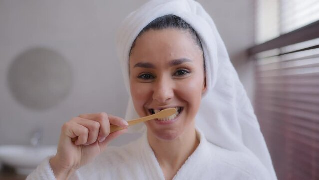 Close-up young woman with towel on head after shower brushing healthy white teeth with toothpaste brushing mouth with toothbrush in home bathroom morning routine dental care personal hygiene concept