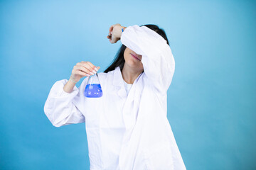 Young brunette woman wearing scientist uniform holding test tube over isolated blue background covering eyes with arm smiling cheerful and funny. Blind concept.