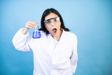 Young brunette woman wearing scientist uniform holding test tube over isolated blue background afraid and shocked with surprise and amazed expression, fear and excited face.