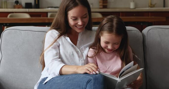 Cheerful young mother and preschool cute daughter having fun at home, spend carefree leisure reading fascinating fairy tale book. Good life habit, intelligence and children development, hobby concept