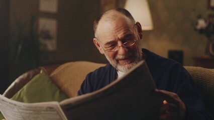 Cheerful senior man sitting on the couch in living room and laughing while watching latest news in the newspaper and reading jokes - Powered by Adobe