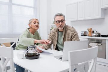 Elderly woman pointing at laptop near husband with paper and coffee in kitchen.