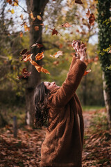 Medium shot of a brunette woman throwing dry leaves into the air in a forest.