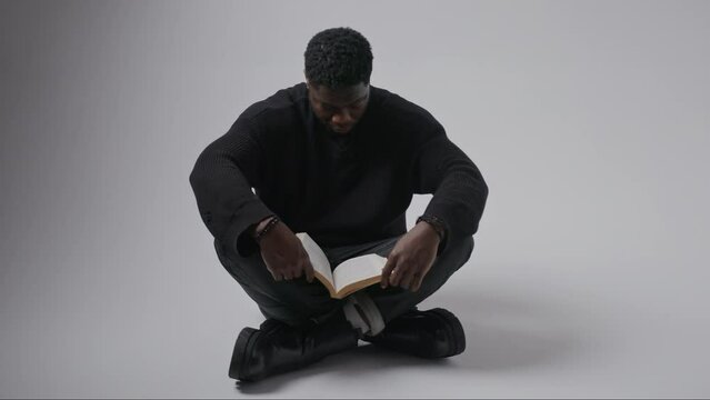 black good looking male reading a book while he's sitting on the floor and nodding his head as a sign of happiness, gray background. High quality 4k footage