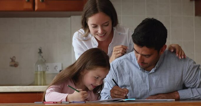 Loving parents their little adorable daughter drawing with colored pencils seated at table in cozy domestic kitchen. Caring mom and dad develop skills of child, enjoy leisure and hobby at home concept