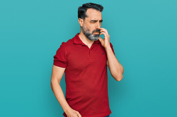 Middle aged man with beard wearing casual red t shirt looking stressed and nervous with hands on mouth biting nails. anxiety problem.