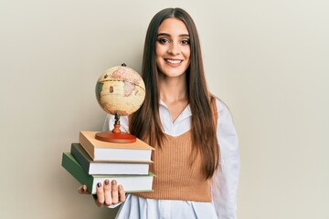 Beautiful brunette young woman holding pile of books and vintage world ball looking positive and...