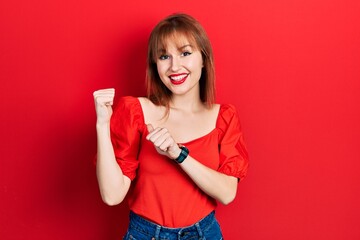 Redhead young woman wearing casual red t shirt pointing to the back behind with hand and thumbs up, smiling confident