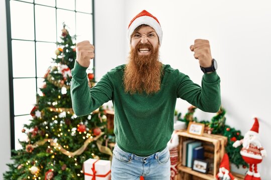 Redhead man with long beard wearing christmas hat by christmas tree angry and mad raising fists frustrated and furious while shouting with anger. rage and aggressive concept.