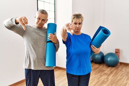 Middle age caucasian couple holding yoga mat at pilates room with angry face, negative sign showing dislike with thumbs down, rejection concept