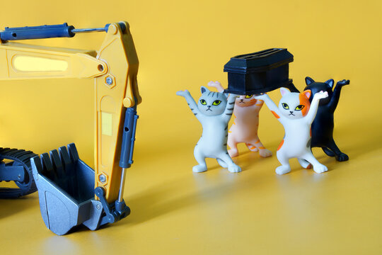 Funny toy kittens carry a black coffin next to the excavator bucket. The concept of a funeral procession dancing with a coffin. Yellow background. Black humor