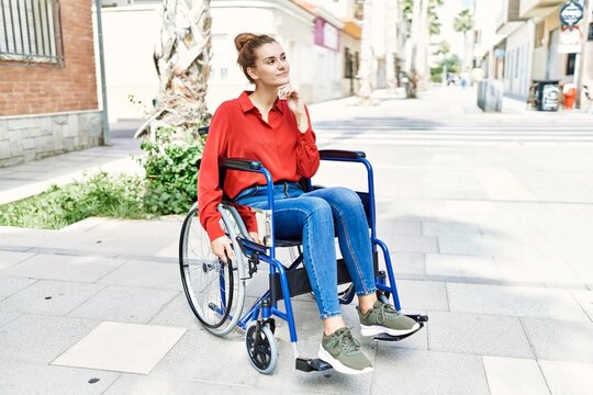 Young brunette woman sitting on wheelchair outdoors serious face thinking about question with hand on chin, thoughtful about confusing idea