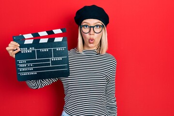 Beautiful blonde woman holding video film clapboard scared and amazed with open mouth for surprise,...