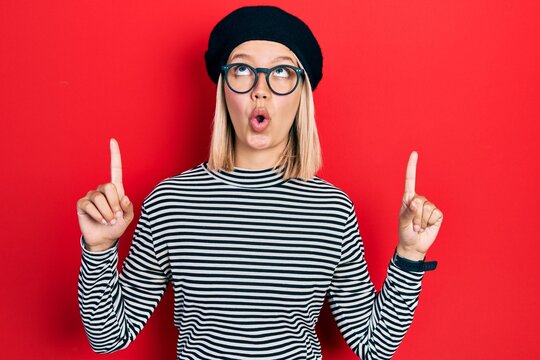Beautiful blonde woman wearing french look with beret an glasses amazed and surprised looking up and pointing with fingers and raised arms.