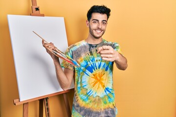 Young hispanic man standing drawing with palette by painter easel stand pointing fingers to camera with happy and funny face. good energy and vibes.