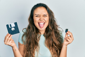 Young hispanic girl holding floppy disk and sdxc card sticking tongue out happy with funny expression.