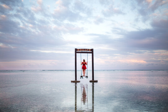 Back view of woman in red dress standing on swinging chair installed on seashore at sunset