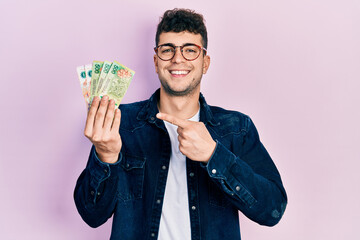 Young hispanic man holding argentine pesos banknotes smiling happy pointing with hand and finger