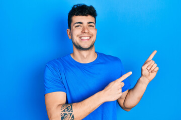 Young hispanic man wearing casual blue t shirt smiling and looking at the camera pointing with two hands and fingers to the side.