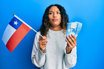 Young latin woman holding chile flag and chilean pesos banknotes clueless and confused expression. doubt concept.