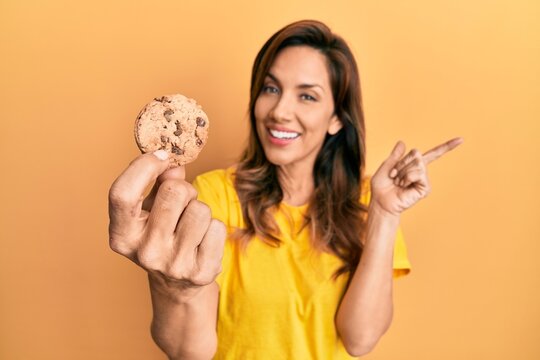 Young latin woman holding cookie smiling happy pointing with hand and finger to the side