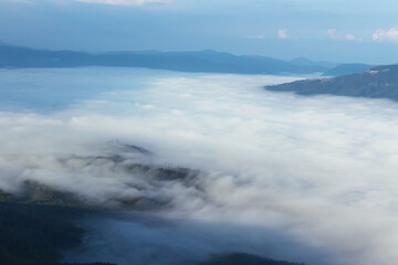 A bird's-eye view of the mountains shrouded in thick fog. In the background are dark silhouettes of ridges and a cloudy sky.