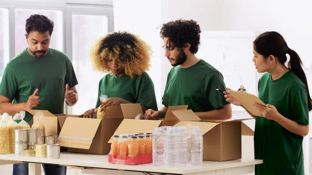 charity, donation and volunteering concept - international group of happy smiling volunteers packing food in boxes according to list on clipboard at distribution or refugee assistance center