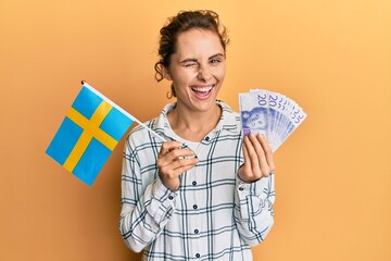 Young brunette woman holding sweden flag and krone banknotes winking looking at the camera with...