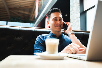 Young smiling handsome man talking at the mobile phone while using computer laptop sitting at cafe table.