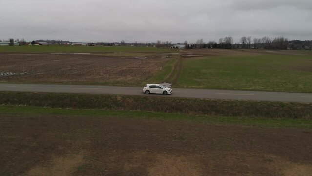 white car 2019 toyota corolla hatchback driving along country farm road surrounded by grass brown fields mountains in farmland Abbotsford BC wide Aerial side profile tracking as car accelerates