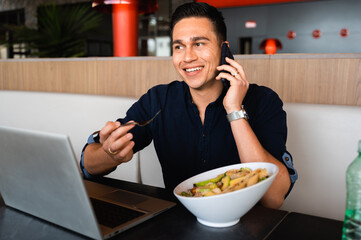 Handsome guy talking at mobile phone with smiling face while eating salad sitting at desk using computer laptop