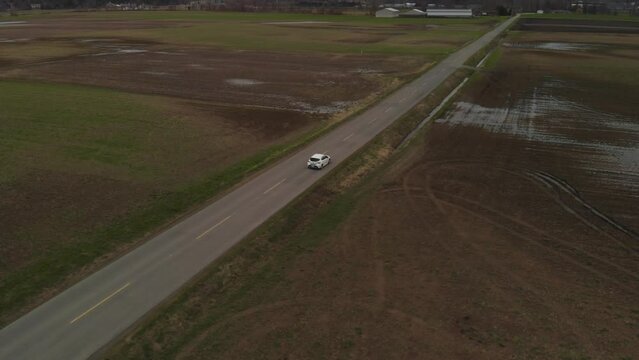 White Car 2019 Toyota Corolla Hatchback Driving Along Country Farm Road Surrounded By Grass Brown Fields Mountains In Farmland Abbotsford BC Aerial Wide Tracking Orbit From Behind Following