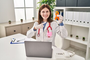 Young doctor woman holding awareness orange ribbon smiling happy pointing with hand and finger