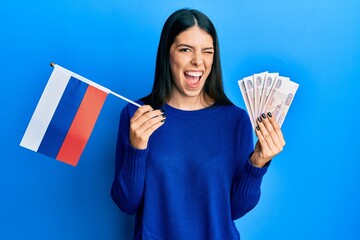 Young hispanic woman holding russia flag and rubles banknotes winking looking at the camera with...
