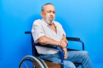 Handsome senior man with beard sitting on wheelchair puffing cheeks with funny face. mouth inflated with air, crazy expression.