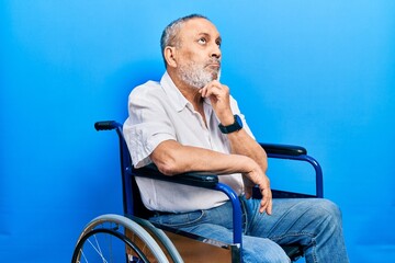 Handsome senior man with beard sitting on wheelchair with hand on chin thinking about question,...