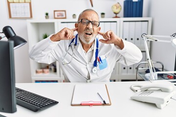 Mature doctor man at the clinic covering ears with fingers with annoyed expression for the noise of loud music. deaf concept.