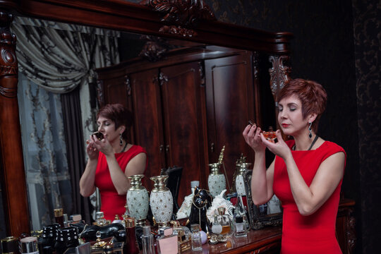 Mature woman in red dress smelling perfume beside dresser