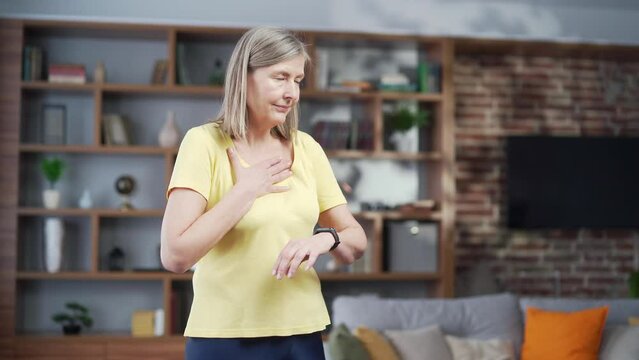 Senior Mature Woman Shortness Of Breath After Workout Looking At Smart Watch Fitness Tracker Bracelet S Holding Chest At Home Indoors. Old Female Increased Heart Rate And Chest Pain