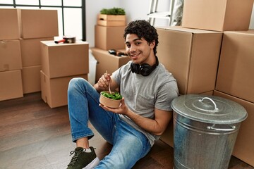 Obraz na płótnie Canvas Young hispanic man eating salad sitting on the floor at new home.