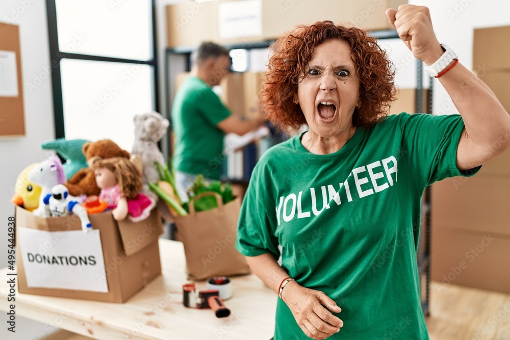 Sticker middle age woman wearing volunteer t shirt at donations stand angry and mad raising fist frustrated 