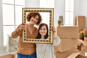 Mature mother and down syndrome daughter moving to a new home, standing by cardboard boxes holding...