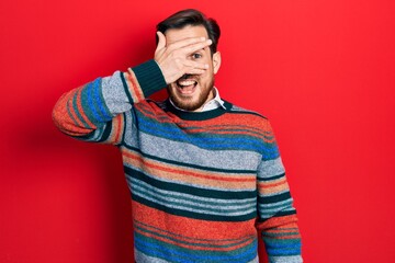 Handsome caucasian man with beard wearing elegant wool winter sweater peeking in shock covering face and eyes with hand, looking through fingers with embarrassed expression.