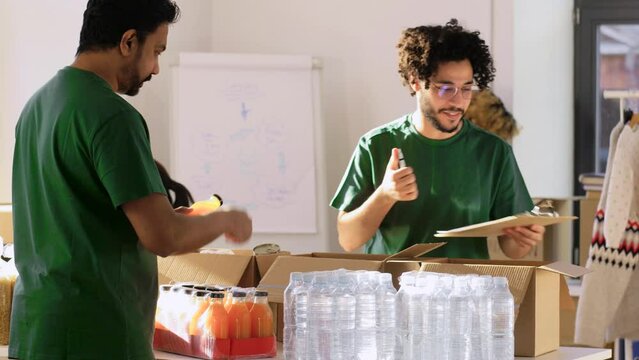 charity, donation and volunteering concept - male volunteers with clipboard packing food and drinks in boxes over international group of people at distribution or refugee assistance center