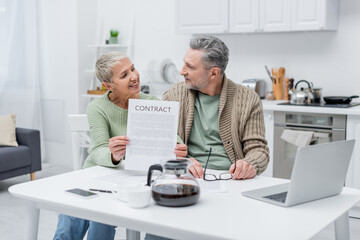 Cheerful senior woman holding contract and looking at husband near gadgets and coffee in kitchen.